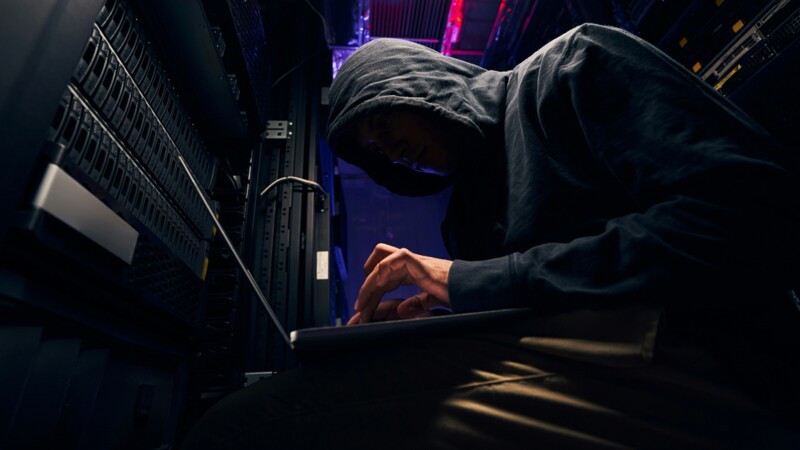 Person mit Kapuzenpullover arbeitet heimlich an Laptop in Serverraum mit dunkler Beleuchtung.