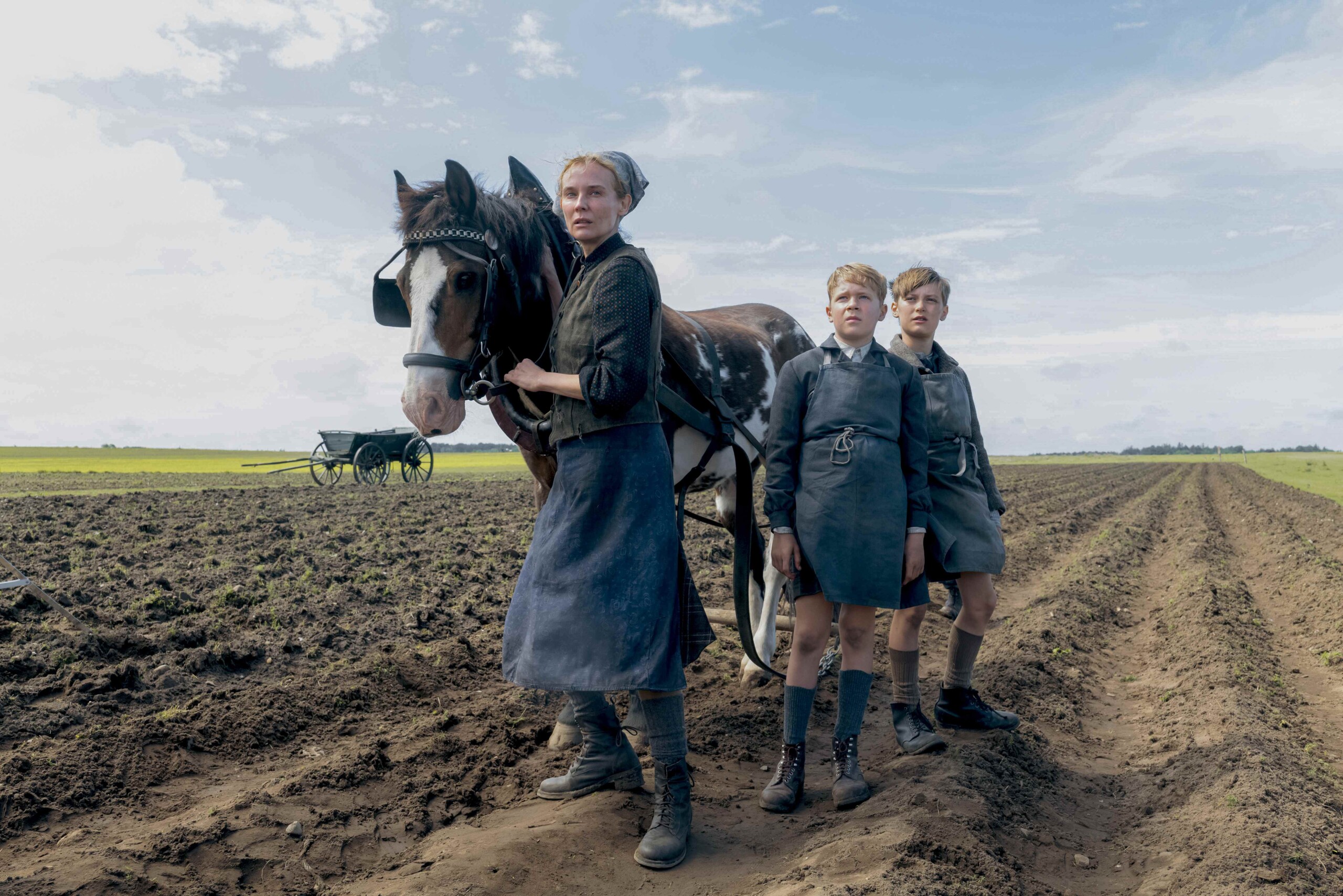 Frau und zwei Jungen stehen neben einem Pferd auf einem gepflügten Feld unter bewölktem Himmel.