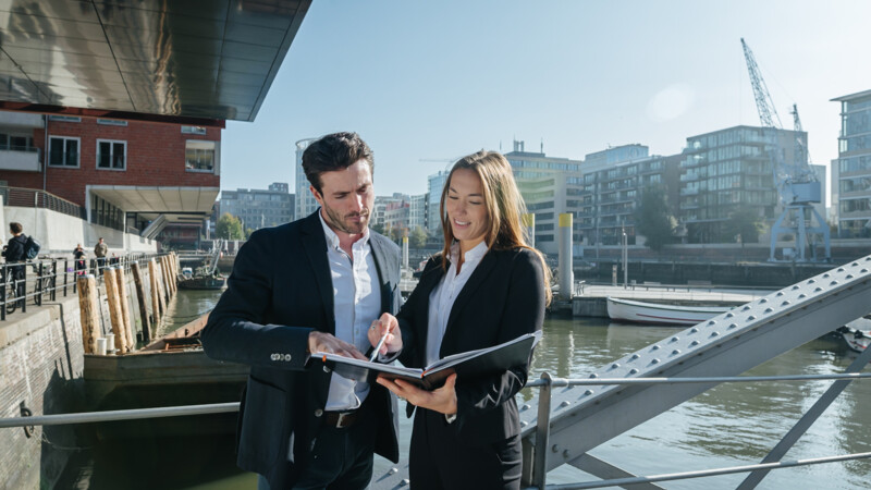 Two business professionals discussing documents on a riverside walkway with modern buildings in the background.