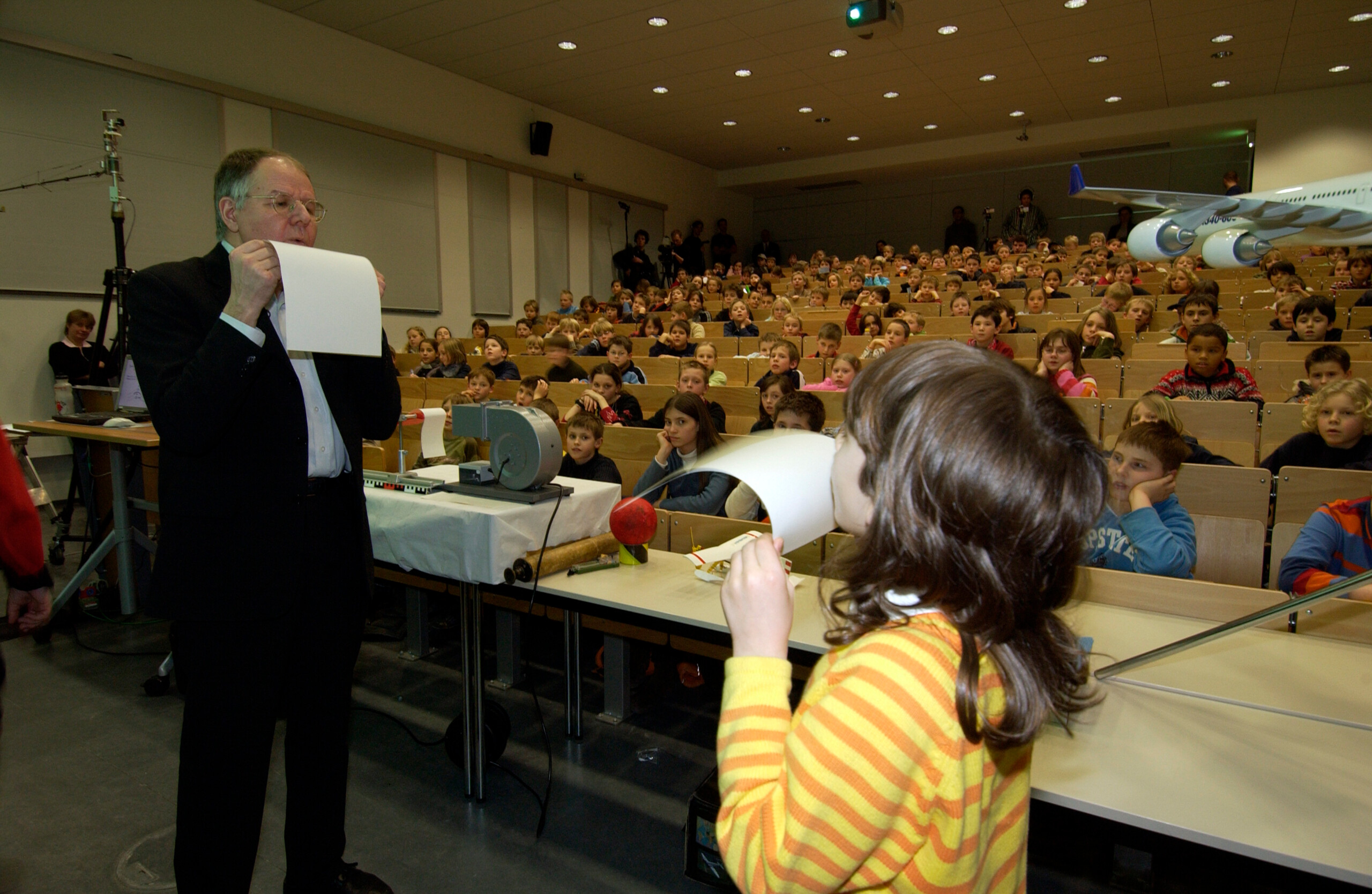 Ein Mann und ein Kind pusten ein Papier an in einem Hörsaal vor einer Gruppe aufmerksam zuhörender Kinder.