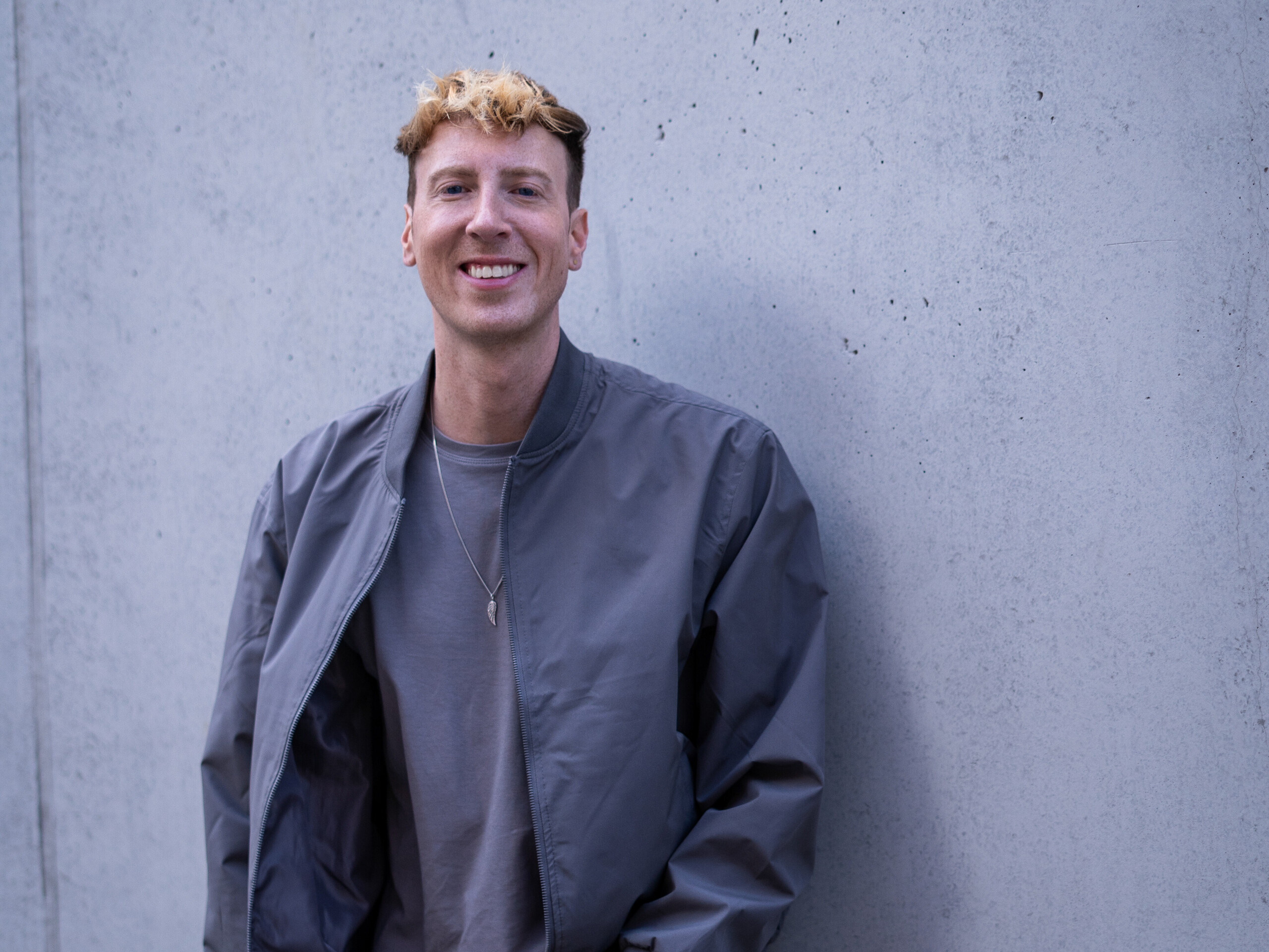 Junger Mann mit blond gefärbten Haaren lächelt vor grauer Betonwand.