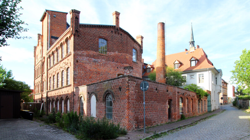 Altes rotes Backsteingebäude mit Schornstein an einer gepflasterten Straße bei klarem Himmel.