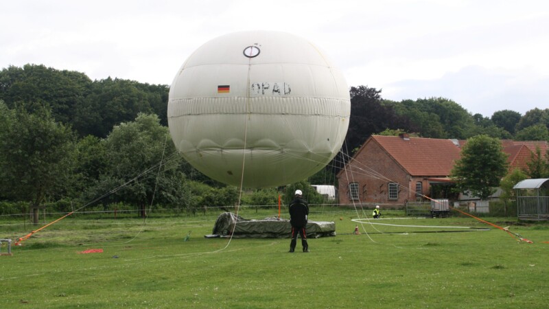 Großer weißer Ballon auf einer Wiese, umgeben von Bäumen und Gebäuden.