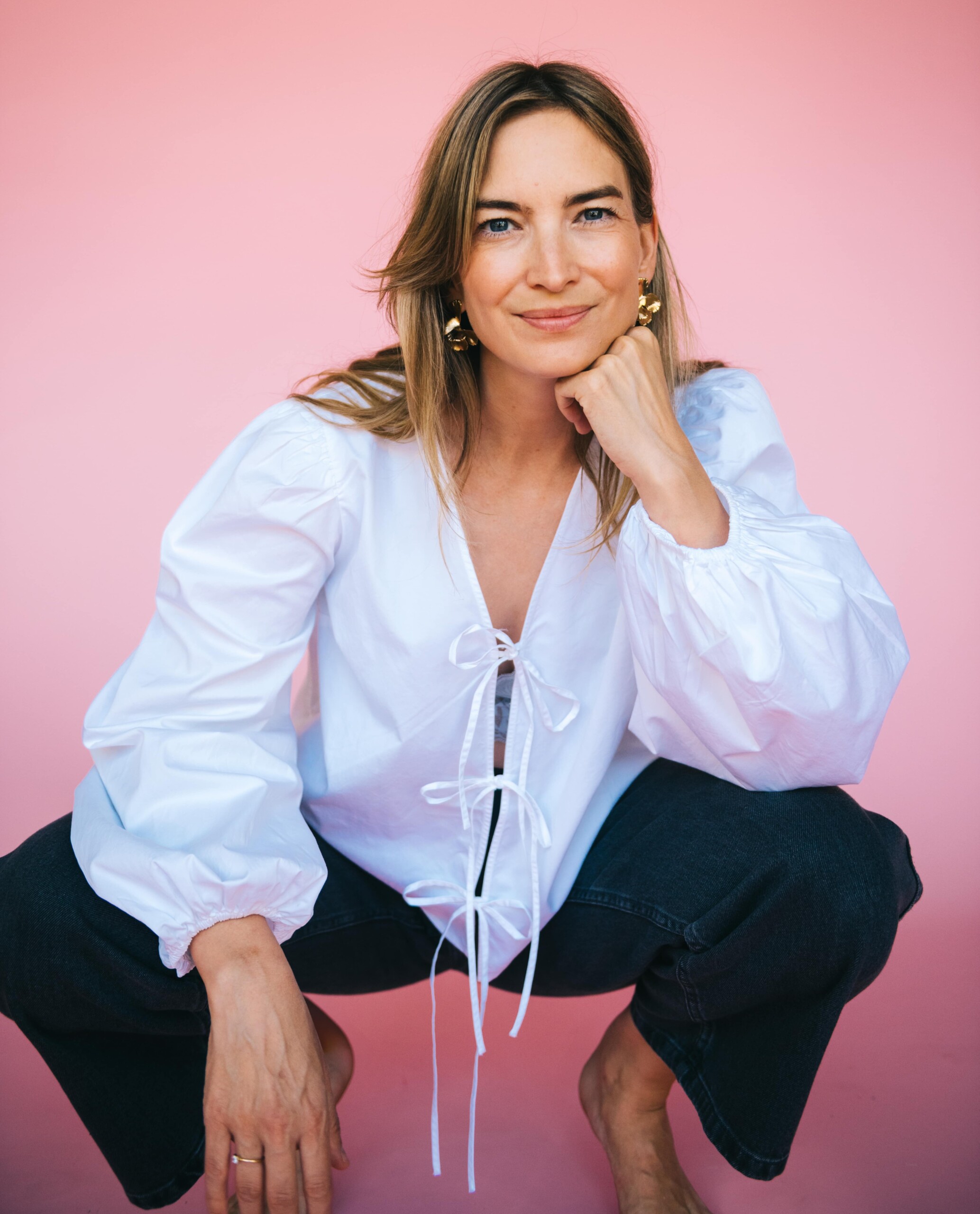 Woman with white blouse and dark trousers in front of a pink background 
