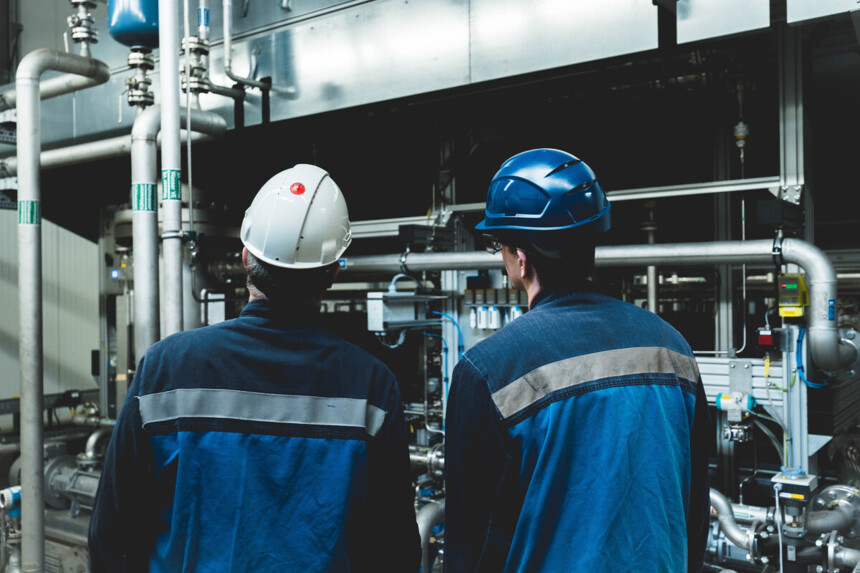 Zwei Industriearbeiter mit Schutzhelmen inspizieren Rohrleitungen in einer Fabrikhalle.