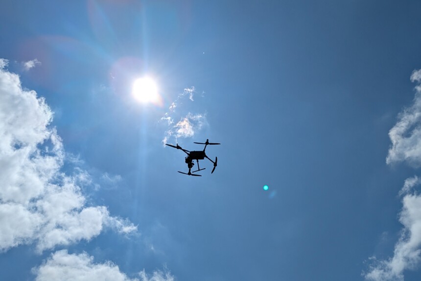 Drohne fliegt vor strahlend blauem Himmel mit Sonne und vereinzelten Wolken.