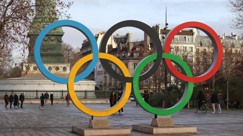Große bunte Olympische Ringe auf einem Platz mit Passanten und historischen Gebäuden im Hintergrund.
