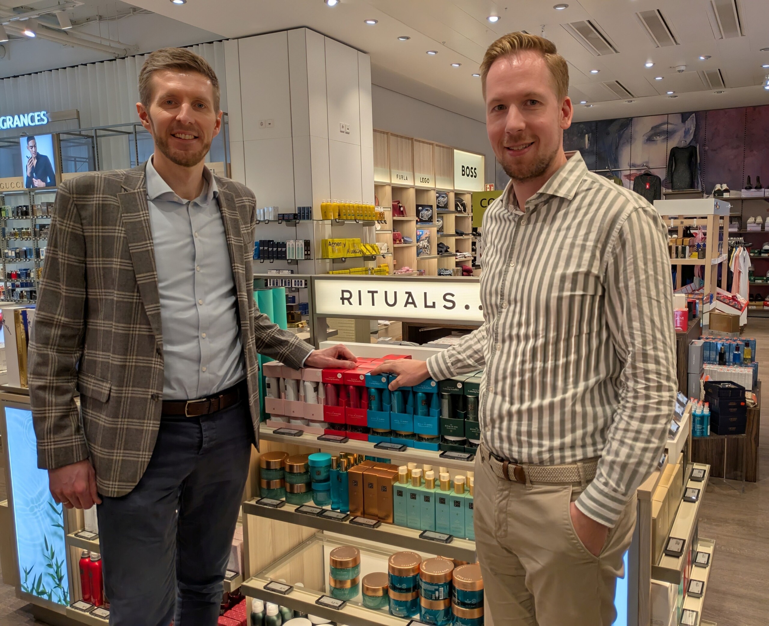 Two men stand smiling in front of a Rituals cosmetics display