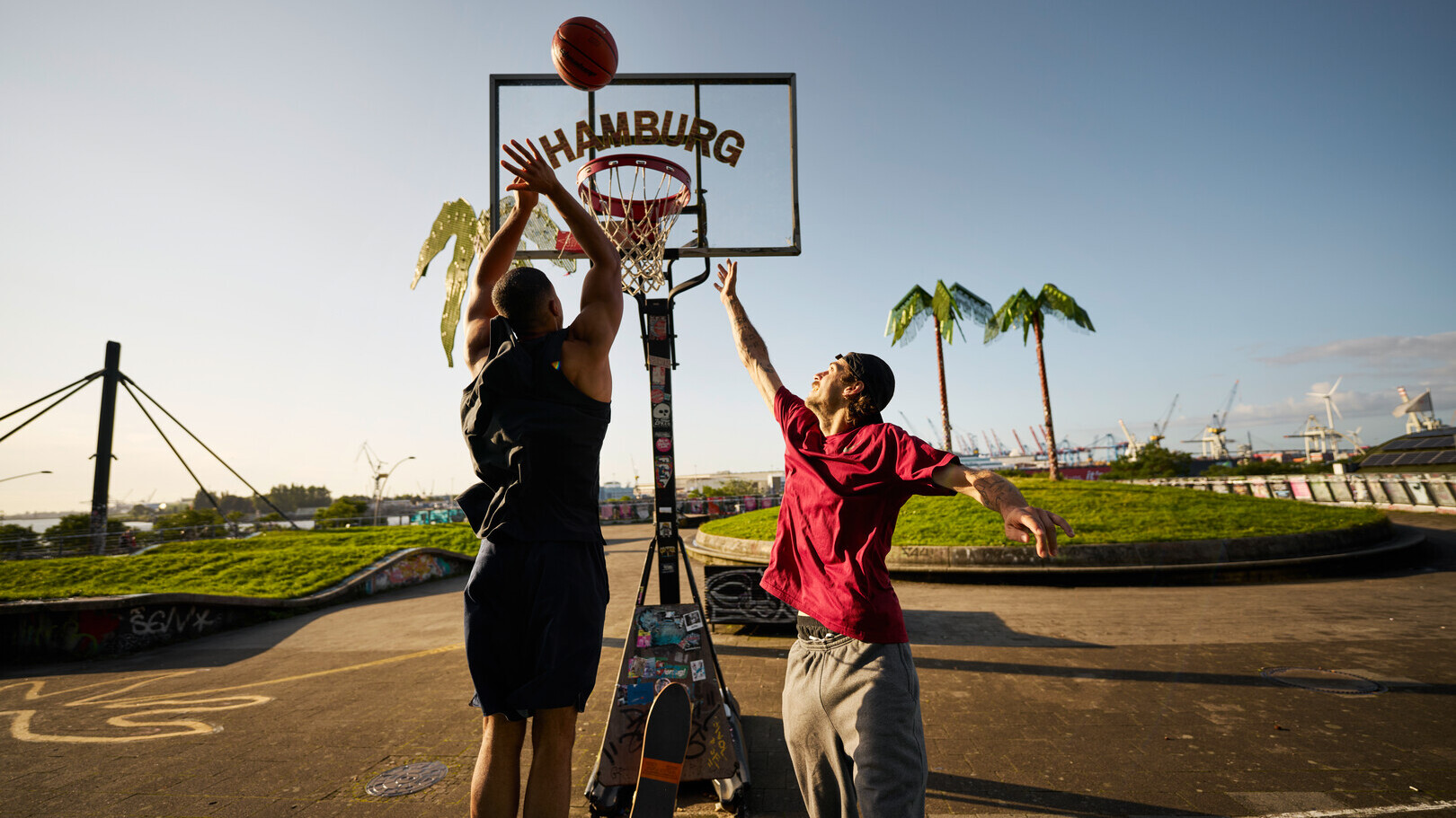 Zwei Männer spielen Basketball auf einem Outdoor-Platz mit Hamburg-Schriftzug und Hafen im Hintergrund.