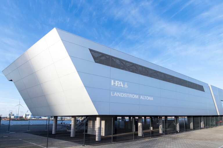 Modernes Gebäude der Hamburg Port Authority mit Beschriftung „Landstrom Altona“ am Hafen unter blauem Himmel.