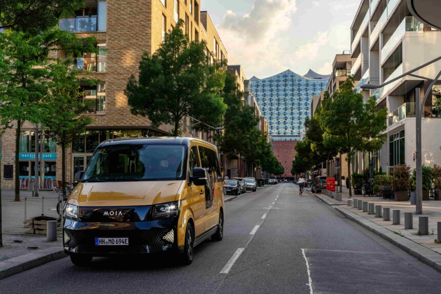 Gelb-schwarzer MOIA-Shuttlebus auf Straße vor moderner Architektur und Bäumen in der Stadt.