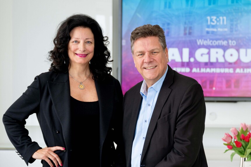 Zwei lächelnde Personen in Businesskleidung vor einem Bildschirm mit Firmenlogo und Blumen auf dem Tisch.