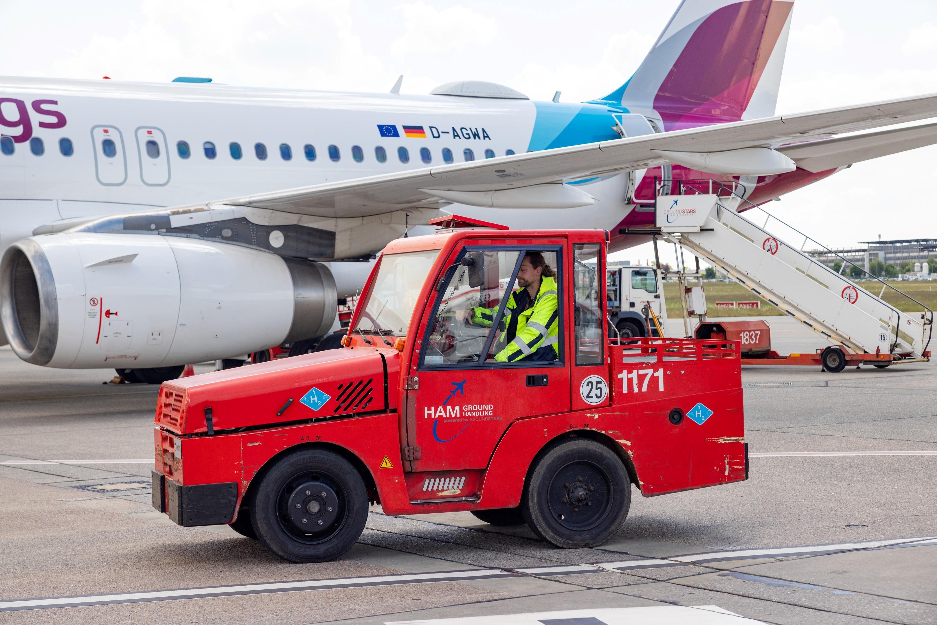 Red hydrogen baggage tug in front of an airplane at Hamburg Airport. The vehicle bears a sticker with the inscription “H2”. An employee in a high-visibility jacket drives the baggage tug