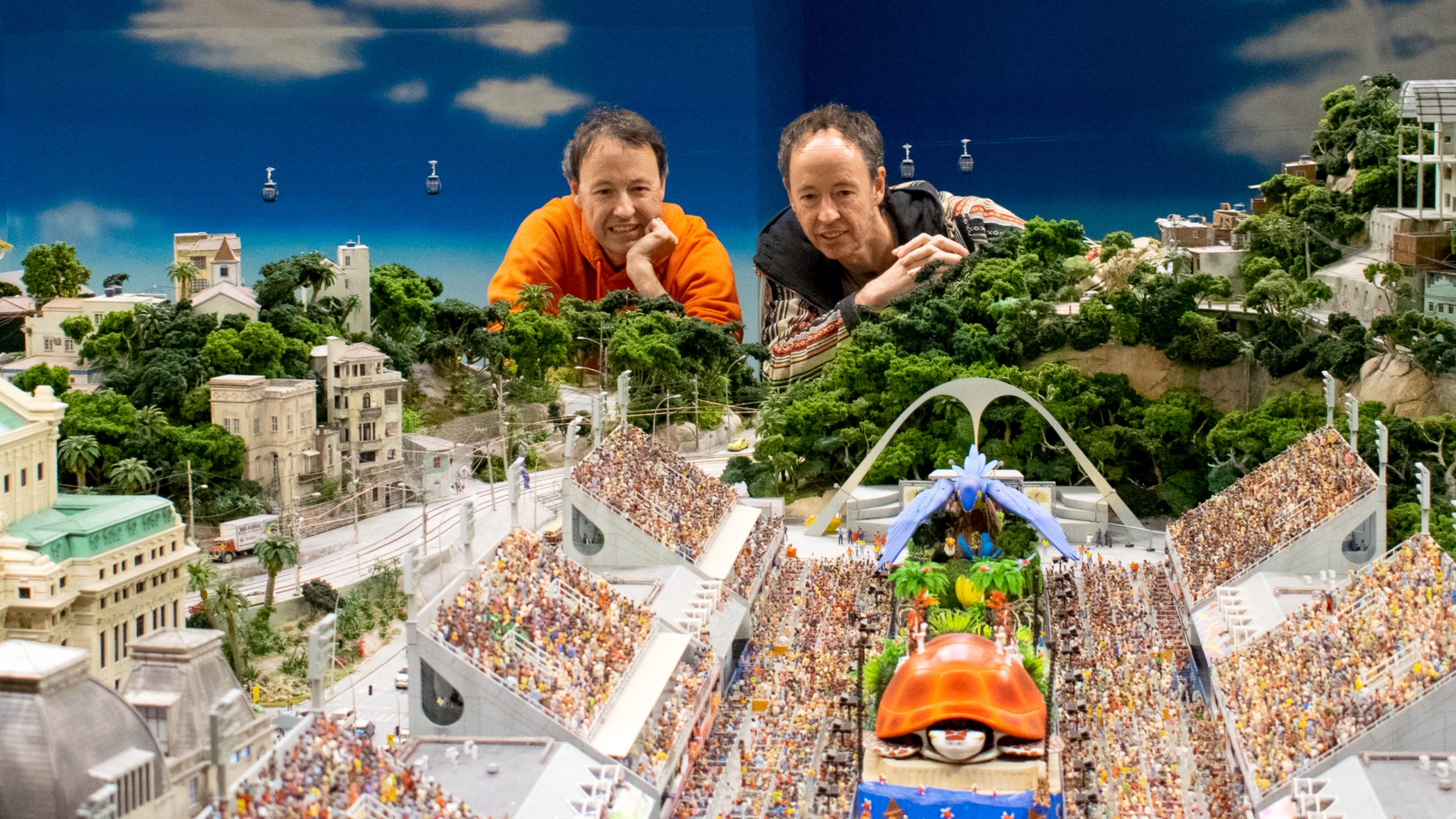 Die Brüder Gerrit und Frederik Braun, Gründer des Miniatur Wunderlands, betrachten ein detailliertes Miniaturmodell eines belebten Karnevalsumzugs mit Zuschauern und grüner Landschaft.