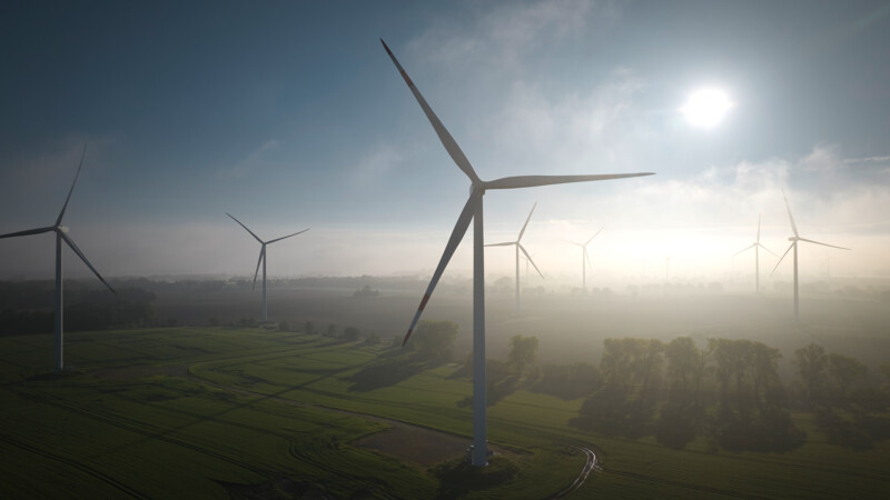 Windkraftanlagen auf grünen Feldern bei Sonnenaufgang mit leichtem Nebel und blauem Himmel.