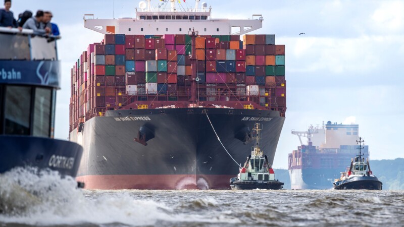 Hafen-Szene: Im Zentrum ein riesiges, von Schleppern gezogenes Containerschiff, links eine Fähre mit Touristen