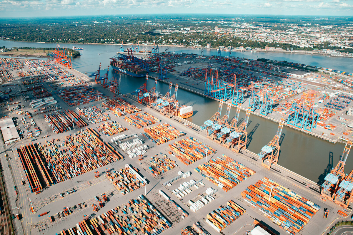 Luftaufnahme eines großen Containerhafens mit zahlreichen Containern, Kränen und einem Frachtschiff am Flussufer.