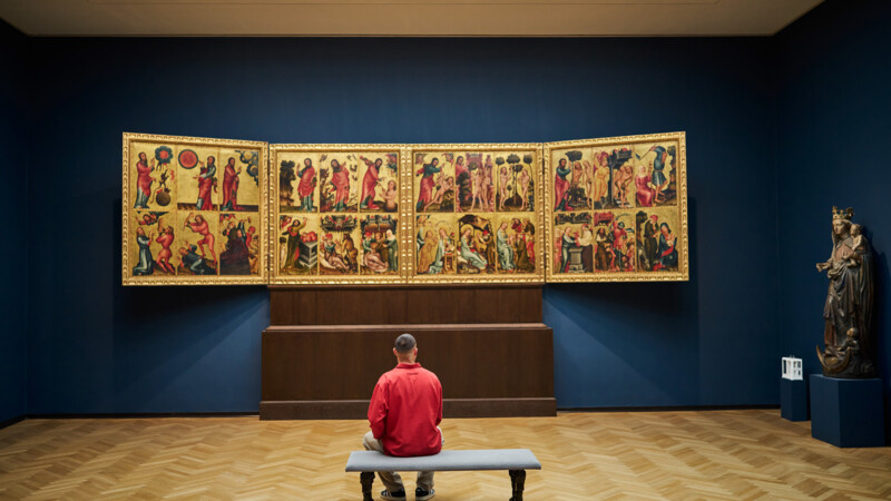 Mann sitzt auf Bank und betrachtet mittelalterlichen Flügelaltar in Museum mit dunklen Wänden.