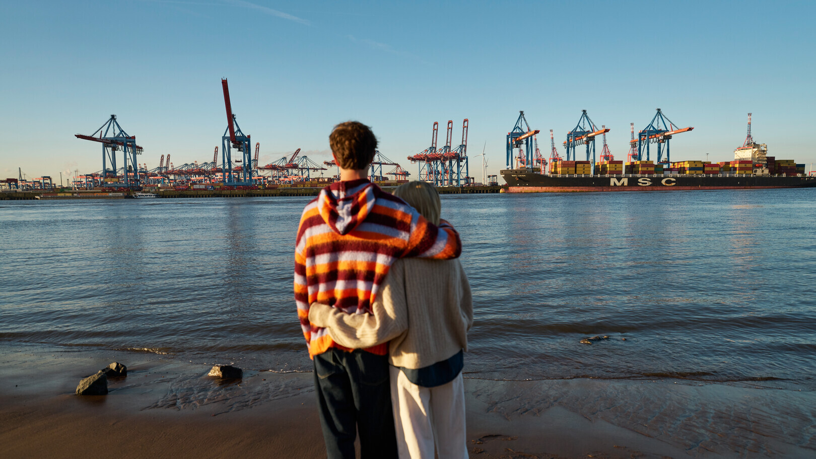 Ein Paar umarmt sich am Ufer und blickt auf einen Containerhafen mit Frachtschiff und Kränen.