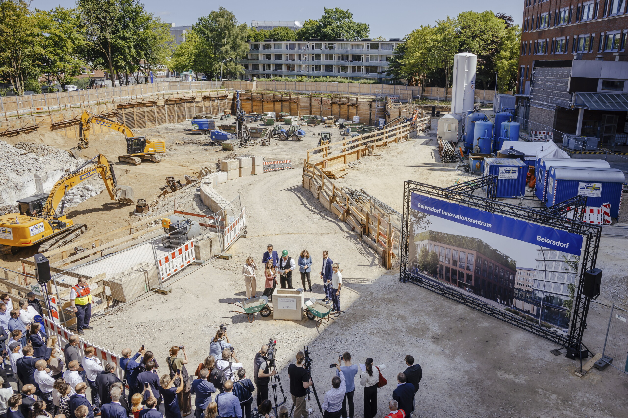 Oblique view from above of the laying of the foundation stone for the innovation centre. In the background excavators and construction site, in the centre the foundation stone with Beiersdorf employees and below journalists and guests observing the event