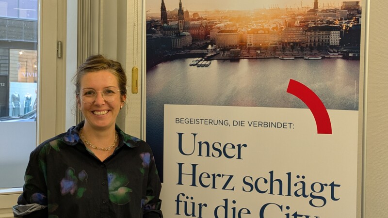 Mimi Sewalske mit Brille lächelt vor einem Plakat mit Hamburg-Panorama und dem Text „Unser Herz schlägt für die City“.
