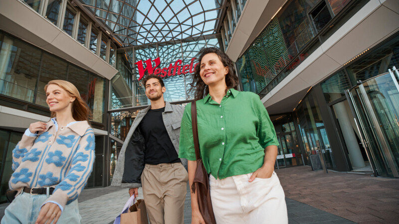 Drei fröhliche Menschen mit Einkaufstüten verlassen das moderne Einkaufszentrum Westfield.
