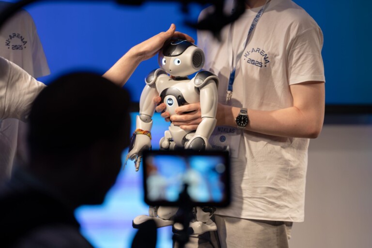 Zwei Personen präsentieren einen NAO-Roboter, während eine Kamera im Vordergrund filmt.