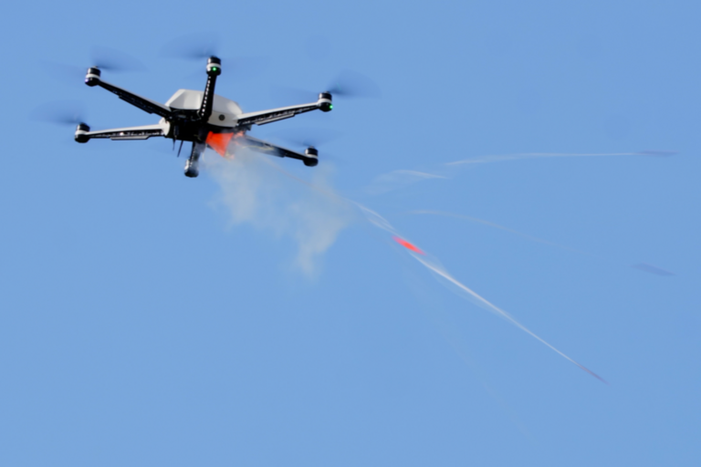 Schwarze Drohne mit sechs Rotoren fliegt am blauen Himmel und stößt Rauch aus.