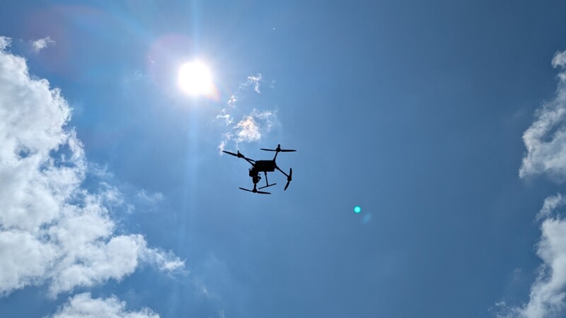 Drohne fliegt vor strahlend blauem Himmel mit Sonne und vereinzelten Wolken.