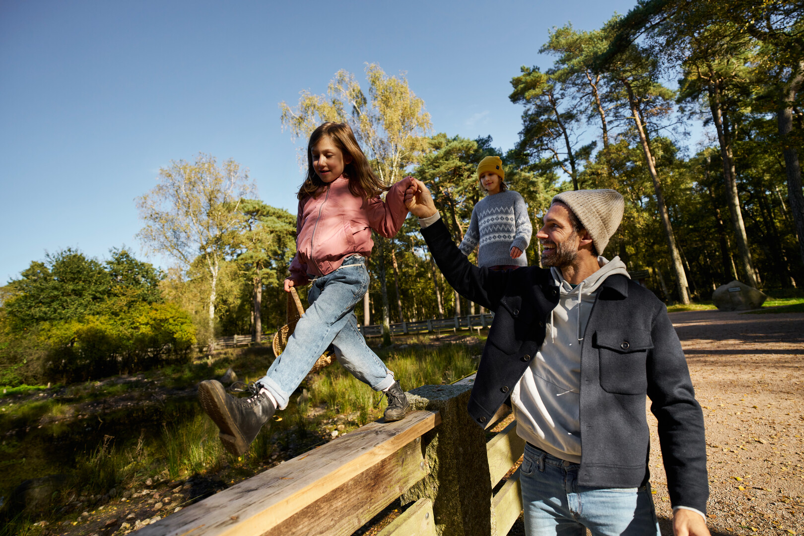 A father helps a girl balance on a wooden fence while another child watches in a sunny forest.