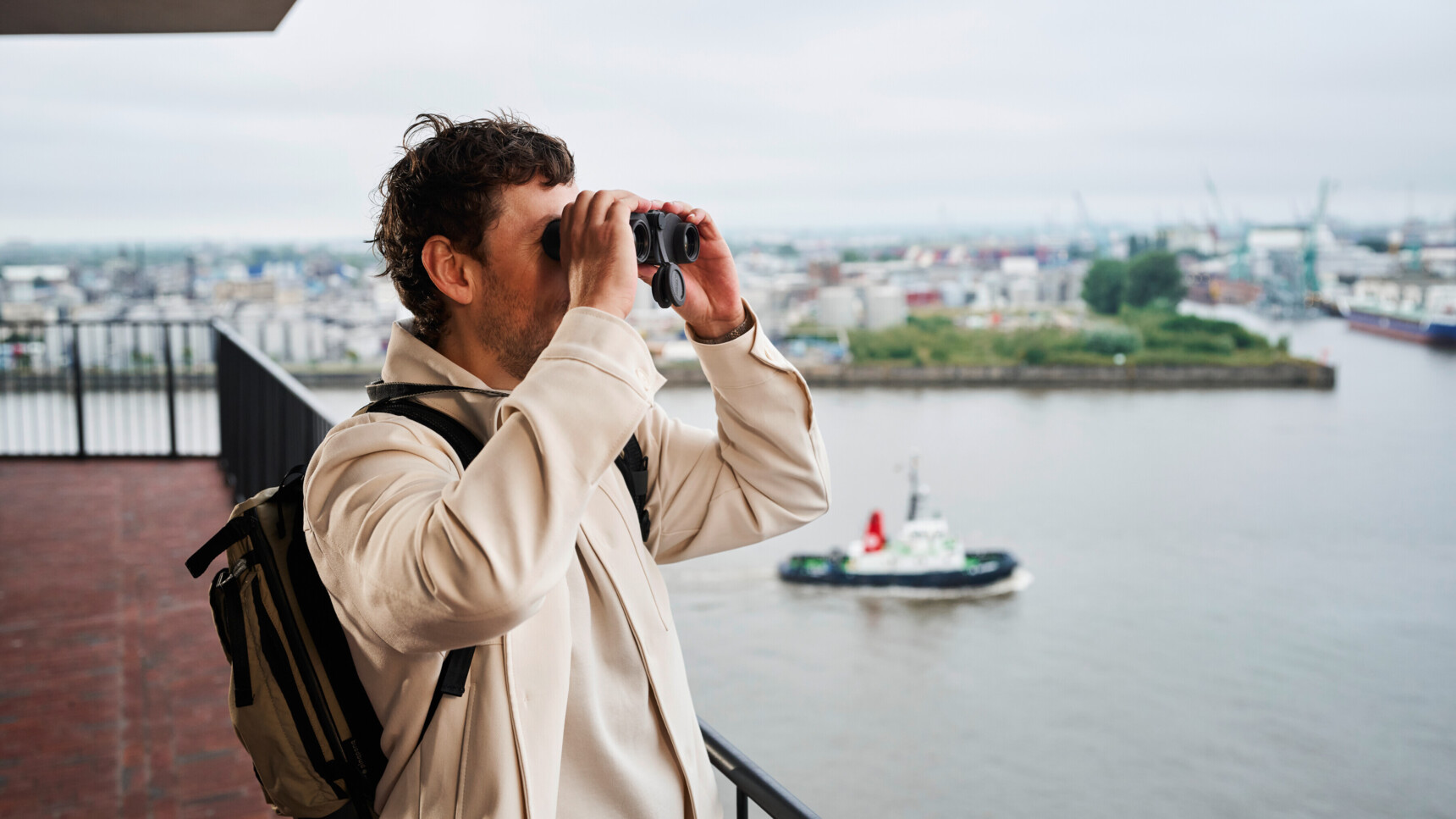 Mann mit Rucksack beobachtet vom Balkon aus mit Fernglas einen Hafen mit Schiffen.