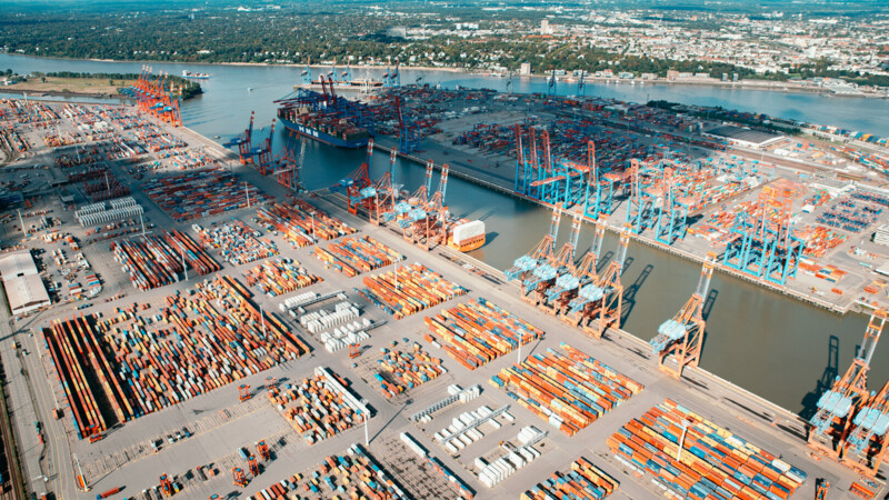 Luftaufnahme eines großen Containerhafens mit zahlreichen Containern, Kränen und einem Frachtschiff am Fluss.