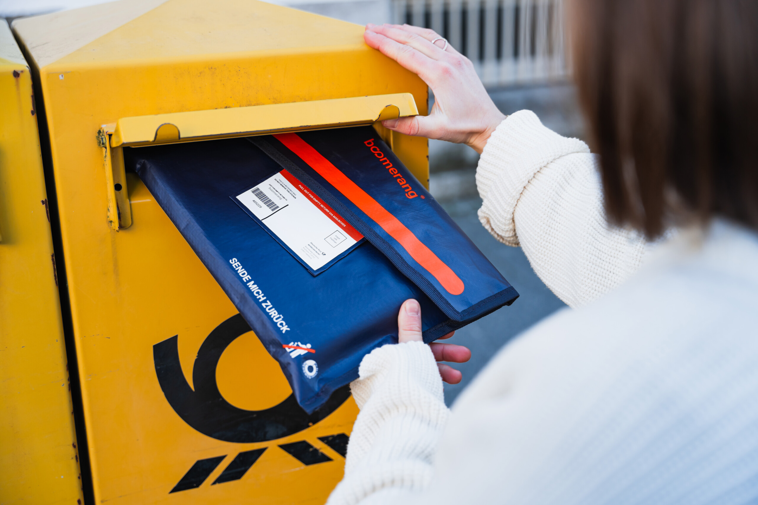 Person wirft blau-rotes Paket in gelben Briefkasten der Deutschen Post ein.