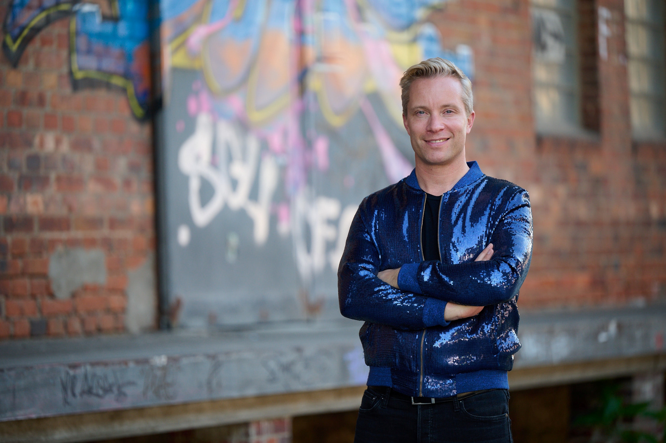 Ulrich Schrauth, Initiator und künstlerischer Leiter von VRHAM!, steht in blauer Glitzerjacke vor einer Wand mit buntem Grafitti