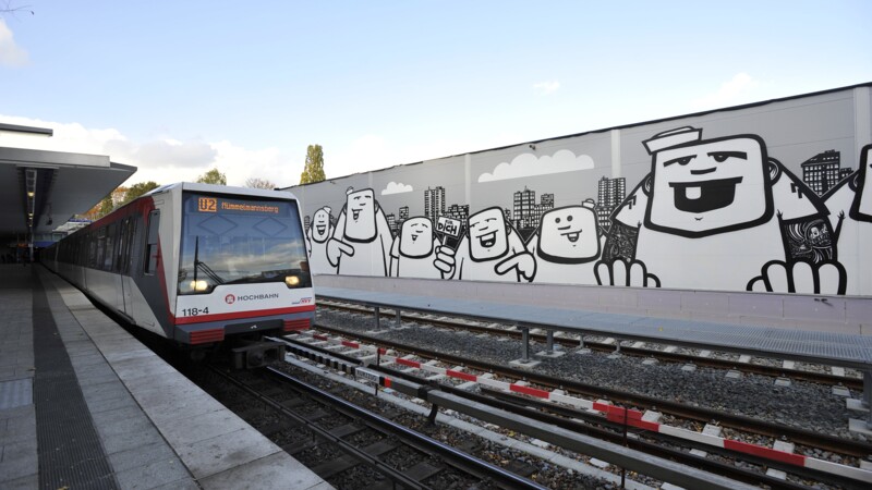 U-Bahn-Zug an einer Station mit großer Wandmalerei von Comicfiguren im Hintergrund.