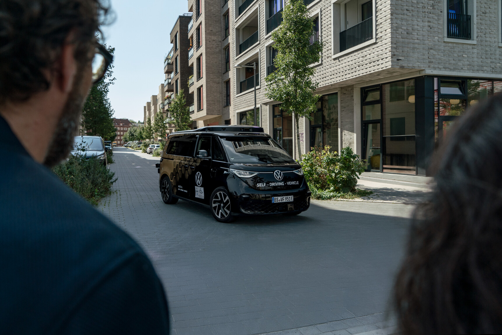 Zwei Personen beobachten einen schwarzen, selbstfahrenden Volkswagen-Bus in einer Wohnstraße.