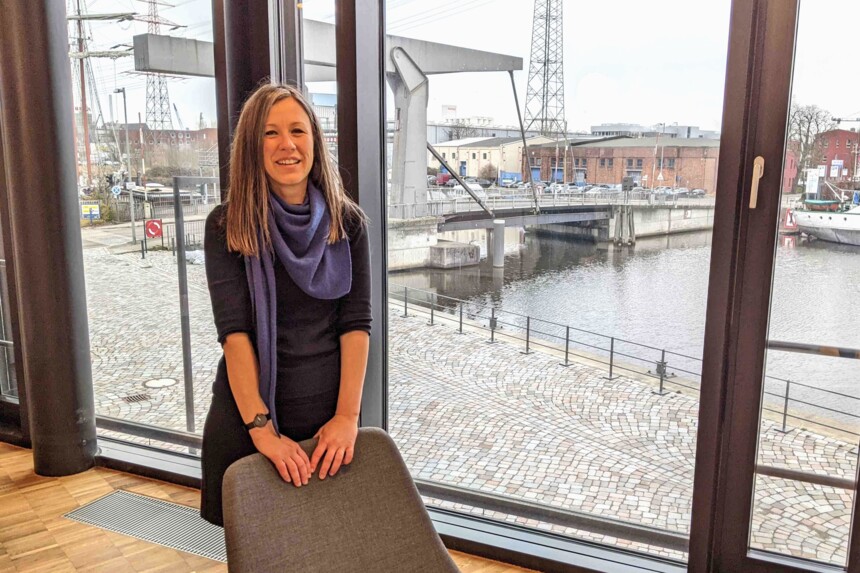 Frau mit blauem Schal steht lächelnd vor einem großen Fenster mit Blick auf einen Fluss und eine Brücke.
