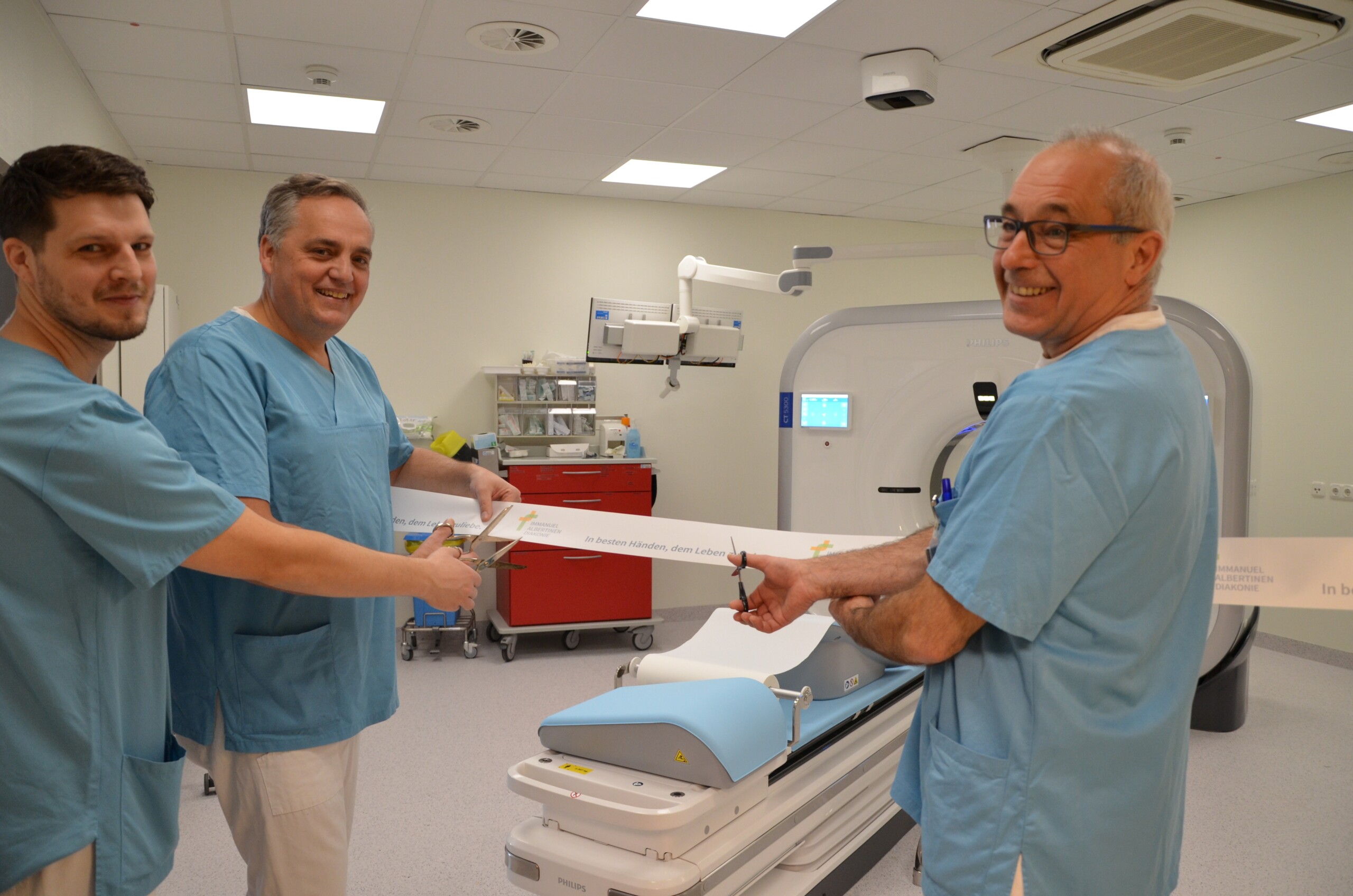 Richard Grundherr, Head Medical Technologist of Radiology, Professor Christoph Weber, Head of Radiology, and Dr Matthias Gasthaus, Head of Cardiology, cut a ribbon in a room with a CT scanner