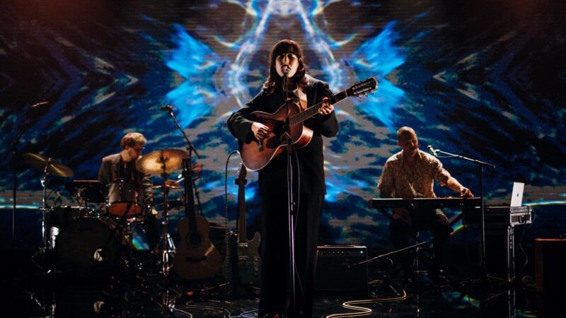 Musician playing guitar and singing with the drummer and keyboarder in front of a blue stage set