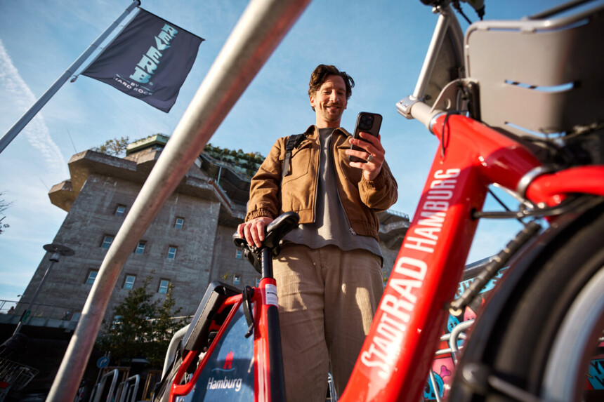 Mann entsperrt ein rotes Stadtrad Hamburg mit seinem Smartphone vor einem Betongebäude.