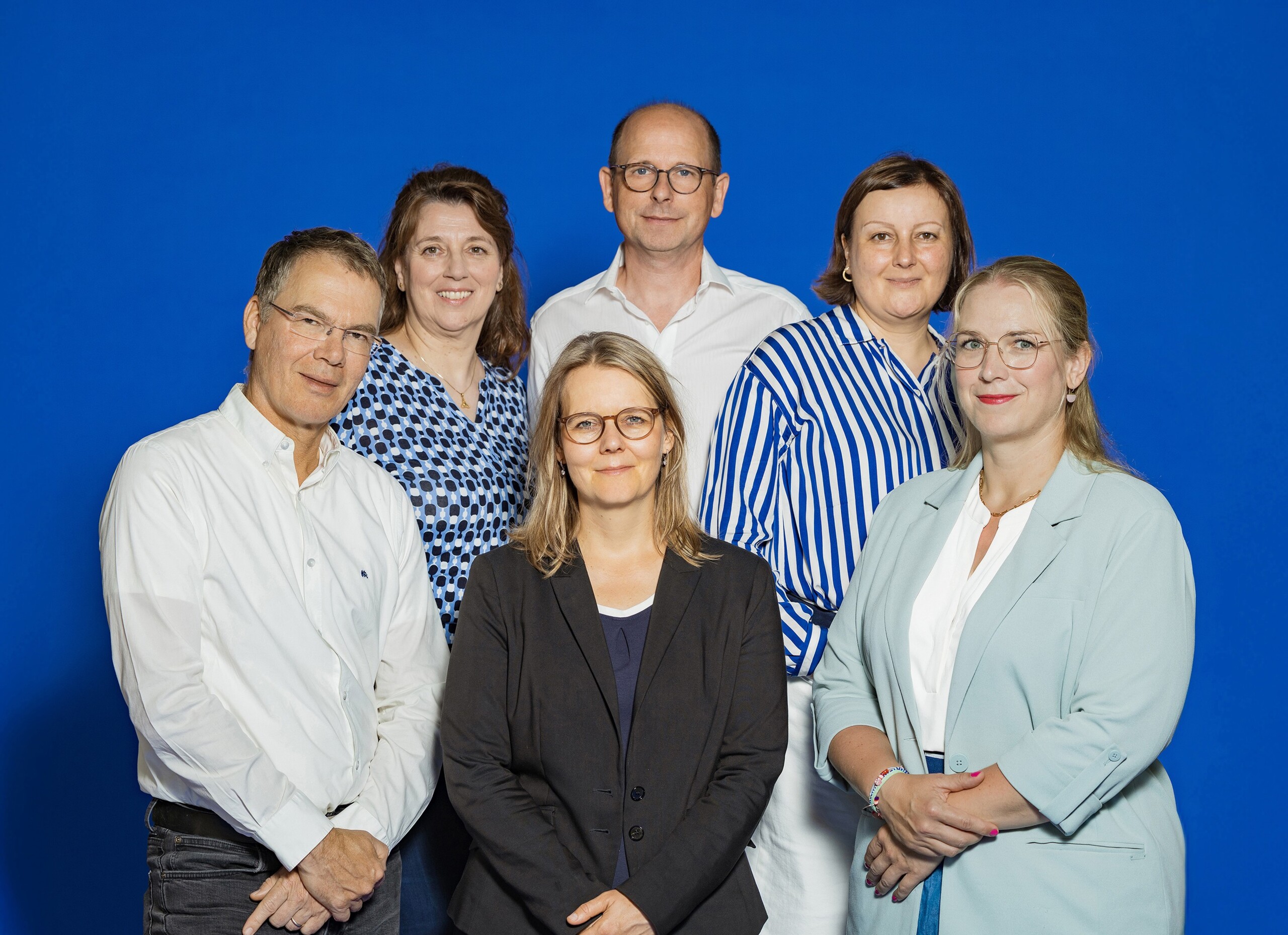 Das Team der Innovations Kontakt Stelle Hamburg: Sechs lächelnde Erwachsene in Business-Kleidung vor blauem Hintergrund, Gruppenfoto.
