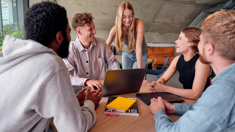 Fünf junge Menschen diskutieren lachend bei einem Meeting um einen Tisch mit Laptop und Notizbüchern.