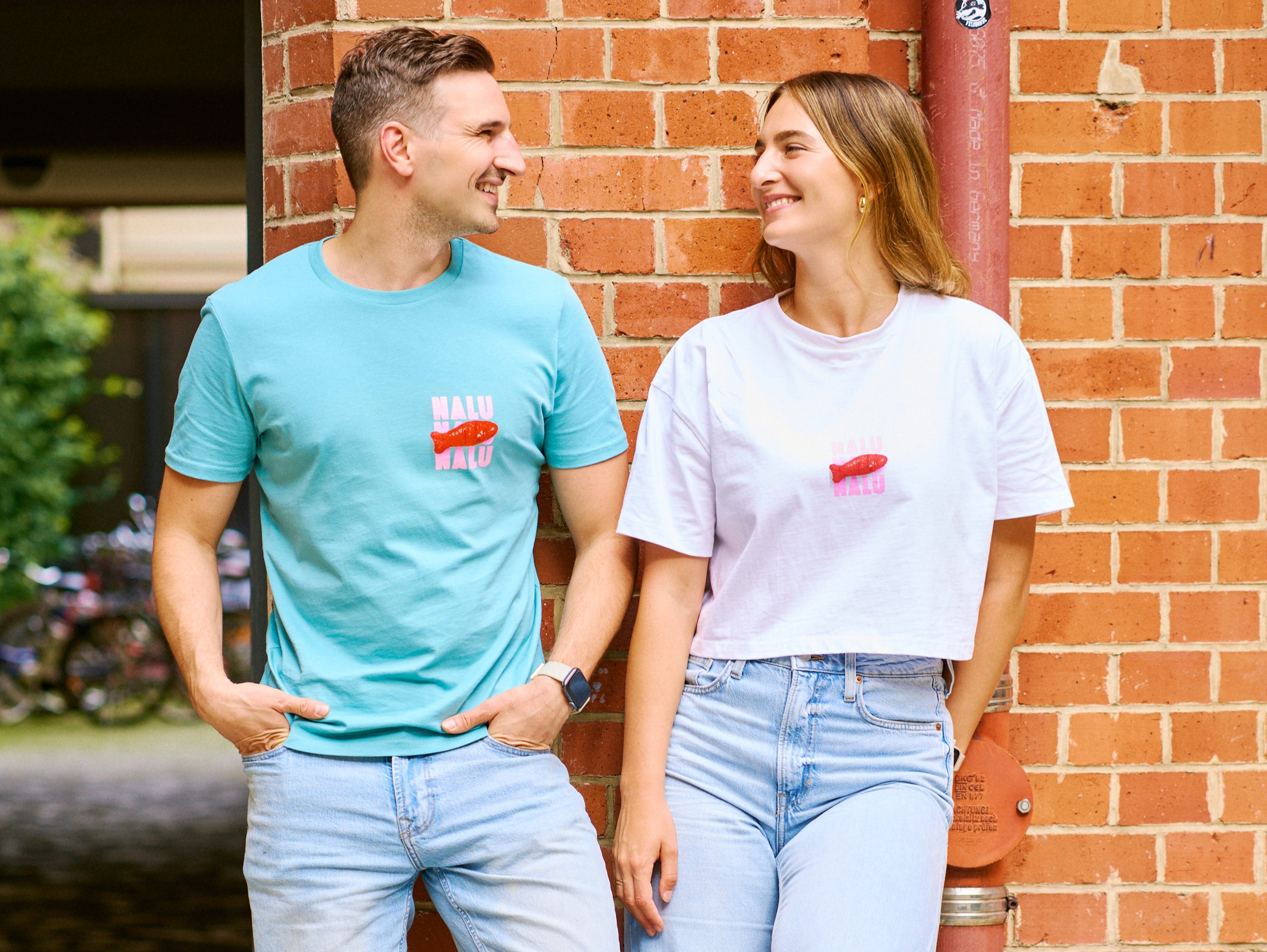 Nalu-Gründer Max Schön und Lisa Ippolito lehnen an einer Backsteinwand und lächeln sich an, beide tragen T-Shirts mit Fischmotiv.