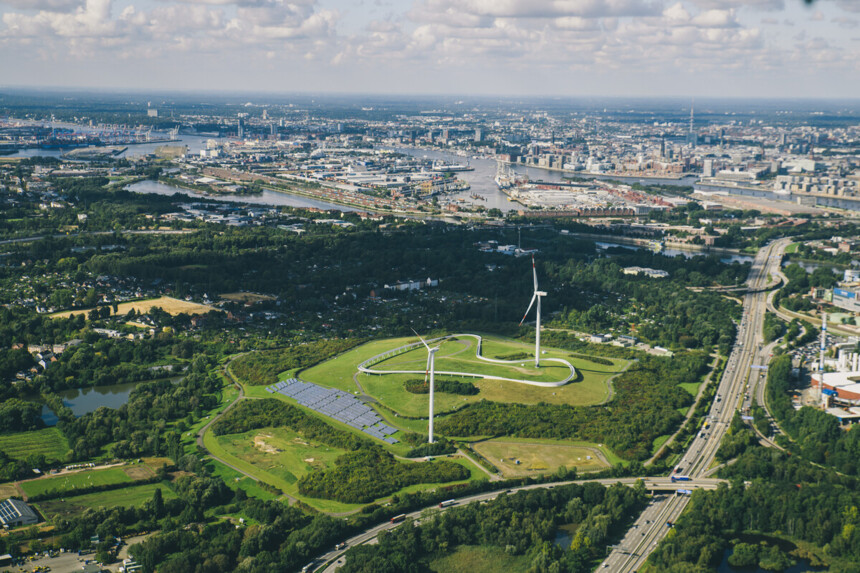 Luftaufnahme von Windrädern und Solarpanelen in einem grünen Park mit Stadt und Hafen im Hintergrund.