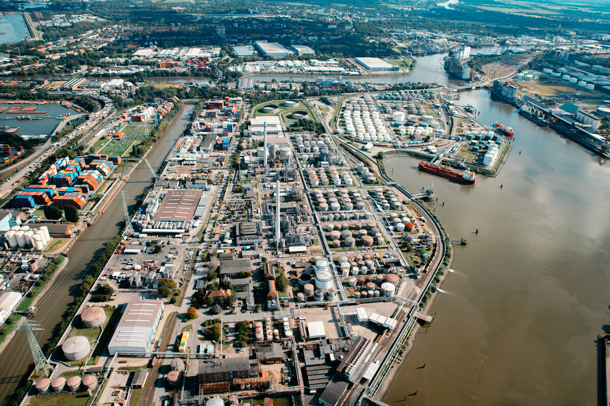 Luftaufnahme eines großen Industriehafens mit Tanks, Lagerhallen und Containerschiffen an einem Fluss.