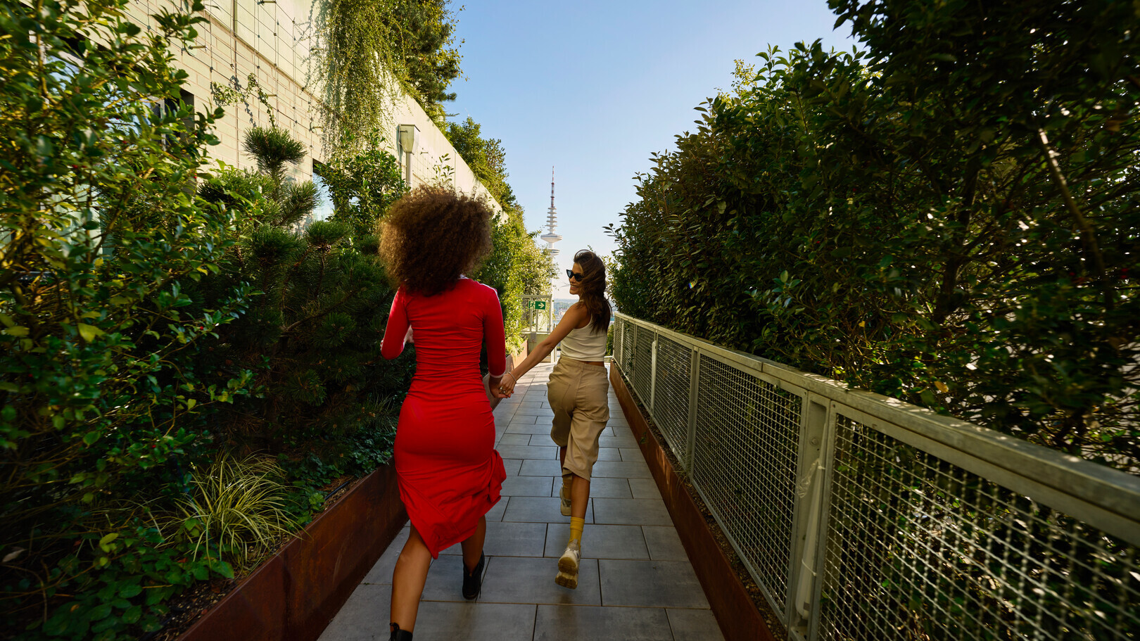 Zwei Frauen laufen Hand in Hand einen begrünten Weg bei sonnigem Wetter entlang.