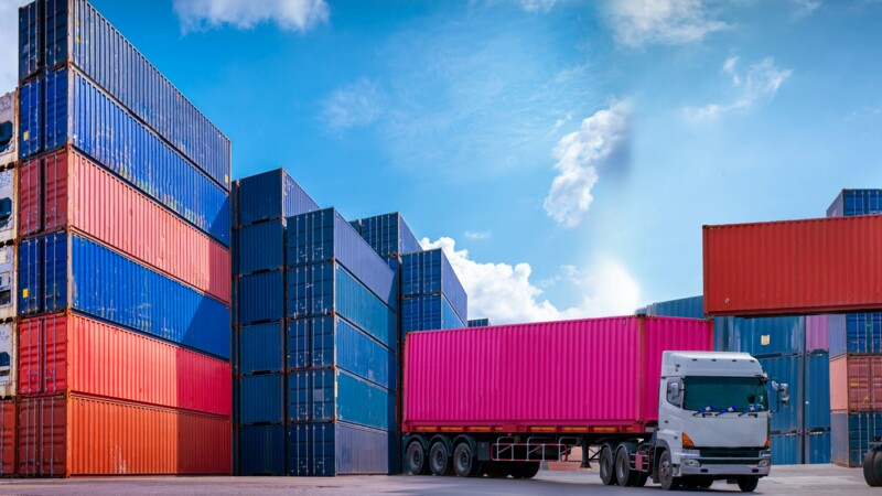 A truck with a pink container is parked among stacked multicoloured shipping containers under a blue sky.