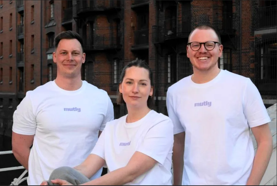 Three people wearing white Mutly t-shirts smiling outside a brick building
